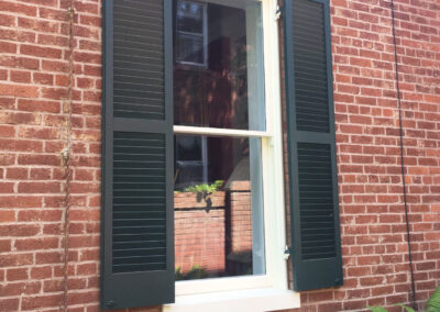 Restored double-hung wood sash window with cream-painted surround and projecting sill, flanked by custom mahogany louvered exterior shutters painted dark green-black. Shutters are full-height louvered stile-and-rail construction, sized correctly to cover the opening if closed — a period-accurate detail often missing in decorative-only installations. Set in historic red brick masonry.